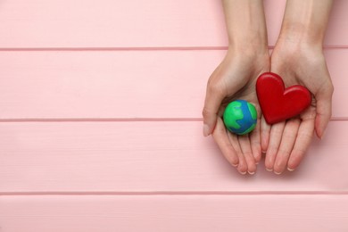 Happy Earth Day. Woman holding plasticine planet and decorative heart at pink wooden table, top view with space for text