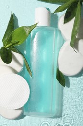 Wet bottle of micellar water, cotton pads and green twigs on light blue background, flat lay