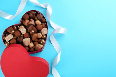 Different delicious chocolate candies in heart shaped box and ribbon on light blue background, top view. Space for text