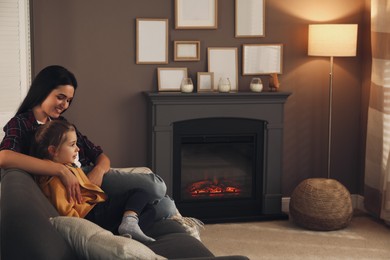 Happy mother and daughter spending time together on sofa near fireplace at home. Space for text