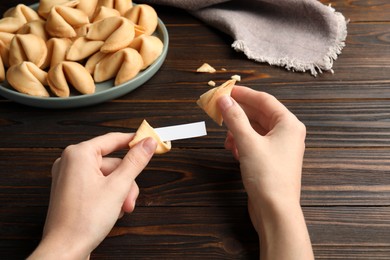 Photo of Woman holding tasty fortune cookie and paper with prediction at wooden table, closeup. Space for text