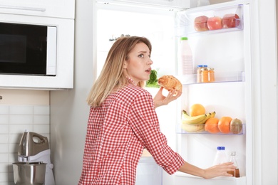 Woman with croissant near fridge in kitchen. Diet failure