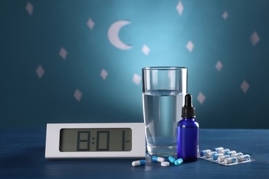Digital alarm clock and different remedies for insomnia treatment near glass of water on table against blue wall decorated with stars and crescent