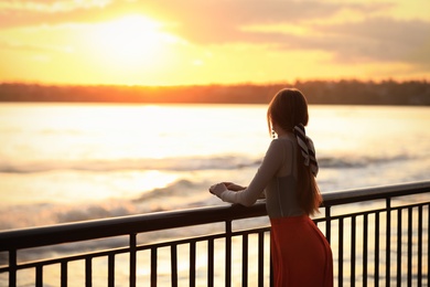 Young woman posing near railing on waterfront at sunset