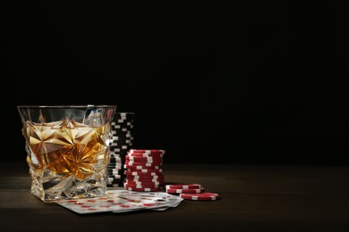 Casino chips, playing cards and glass of whiskey on wooden table, space for text