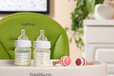 Photo of High chair with feeding bottles of infant formula and toy maracas on white tray indoors