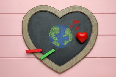 Photo of Happy Earth Day. Blackboard with drawing of planet, chalk pieces and decorative heart on pink wooden table, top view