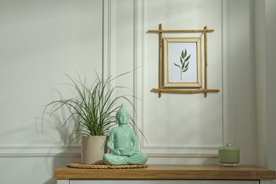 Potted houseplant, buddha statue and candle in jar on wooden table near white wall with stylish bamboo frame