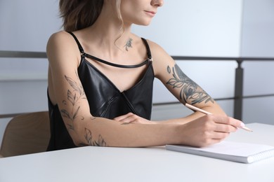 Beautiful woman with tattoos on body drawing in sketchbook at table indoors, closeup