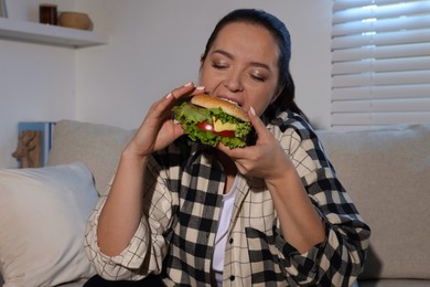 Overweight woman eating burger on sofa at home