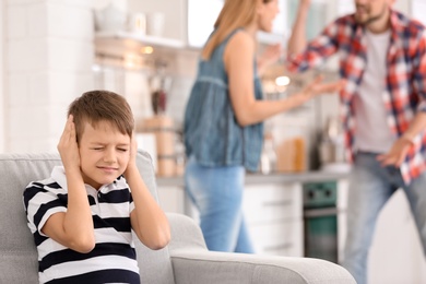Little unhappy boy sitting on sofa while parents arguing at home