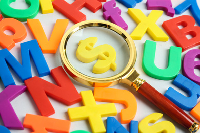 Magnifying glass over dollar sign surrounded by magnet letters on white background, closeup. Search concept