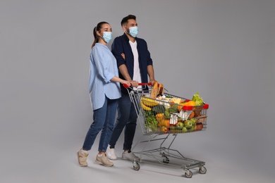 Couple with protective masks and shopping cart full of groceries on light grey background