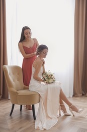 Gorgeous bride in beautiful wedding dress and her friend making hairstyle in room