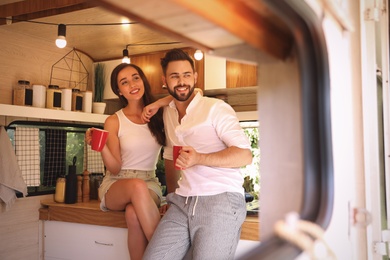Photo of Happy young couple with cups in trailer, view from outside. Camping vacation