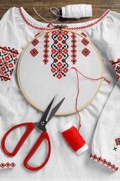 Shirt with red embroidery design in hoop, needle, scissors and threads on wooden table, flat lay. National Ukrainian clothes