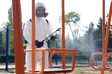 Man in hazmat suit spraying disinfectant onto swing at children's playground. Surface treatment during coronavirus pandemic