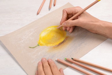 Woman drawing lemon on paper with pastel pencil at white wooden table, closeup