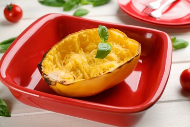 Half of cooked spaghetti squash with basil in baking dish on white wooden table, closeup