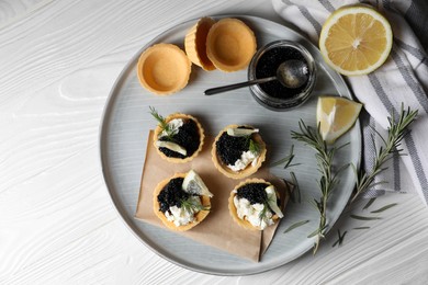 Photo of Plate of delicious tartlets with black caviar, cream cheese and lemon served on white wooden table, top view. Space for text