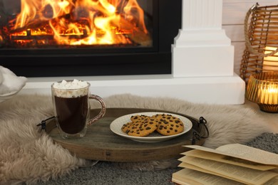 Open book, hot drink and cookies near decorative fireplace in room. Cozy home atmosphere