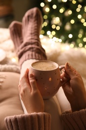 Woman holding cup of delicious hot drink near Christmas tree indoors, closeup