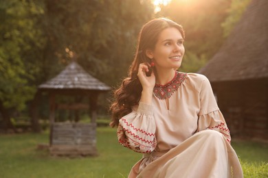 Beautiful woman wearing embroidered dress in village on sunny day, space for text. Ukrainian national clothes
