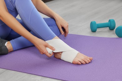 Photo of Woman wrapping foot in medical bandage on yoga mat indoors, closeup