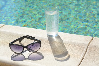Stylish sunglasses and glass of water near outdoor swimming pool on sunny day