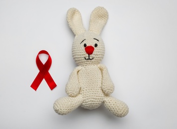 Photo of Cute knitted toy bunny and red ribbon on light grey background, flat lay. AIDS disease awareness