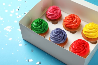Box with delicious colorful cupcakes and confetti on light blue background