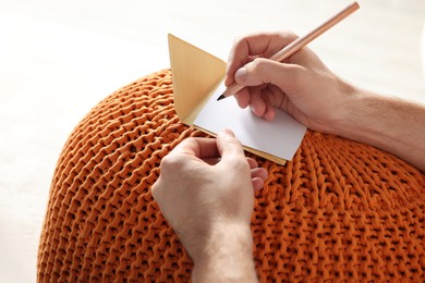 Photo of Man writing message in greeting card on orange
knitted pouf, closeup