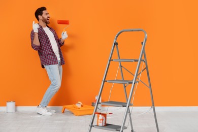 Photo of Emotional designer with roller and painting equipment near freshly painted orange wall indoors