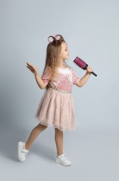 Cute little girl with hairbrush singing on light grey background