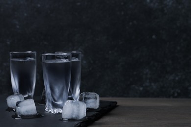 Shot glasses of vodka with ice cubes on wooden table against black wall. Space for text