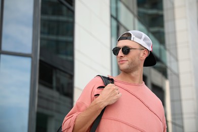 Handsome young man with stylish sunglasses and backpack near building outdoors, space for text