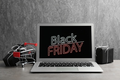 Laptop with Black Friday announcement, small shopping cart and gifts on table against grey background
