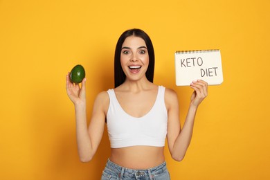 Happy woman holding avocado and notebook with words Keto Diet on yellow background