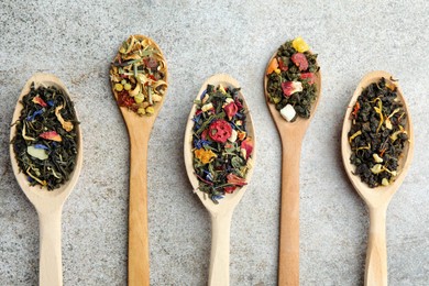Different kinds of dry herbal tea in wooden spoons on light grey table, flat lay