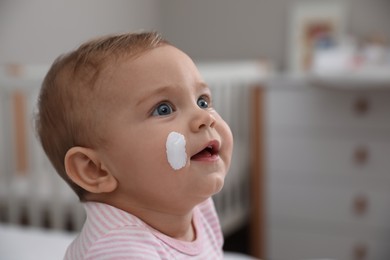 Cute little baby with moisturizing cream on her face at home, closeup. Space for text