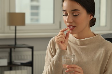 Young woman with glass of water taking dietary supplement pill indoors, space for text