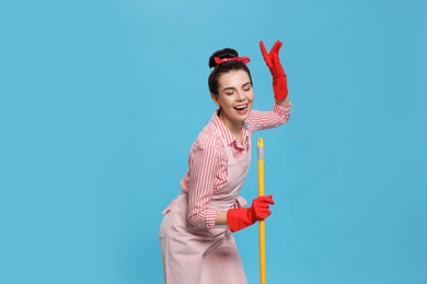 Young housewife with broom having fun on light blue background