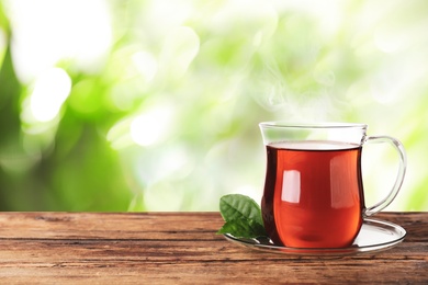 Glass cup of fresh hot tea on wooden table against blurred green background. Space for text
