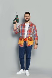 Young worker with power drill and tool belt on grey background