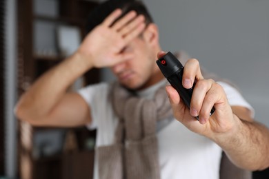 Man covering eyes with hand and using pepper spray indoors, focus on canister