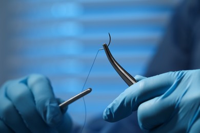 Professional surgeon holding forceps with suture thread on blurred background, closeup. Medical equipment