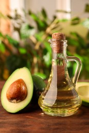 Photo of Glass jug of cooking oil and fresh avocados on wooden table against blurred green background