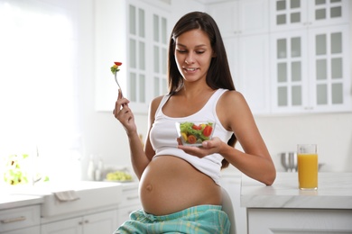 Young pregnant woman with bowl of vegetable salad at table in kitchen. Taking care of baby health