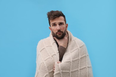 Man wrapped in blanket suffering from sore throat on light blue background. Cold symptoms