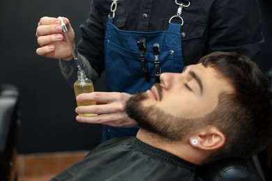Hairdresser with beard oil near client in barbershop, closeup. Professional shaving service